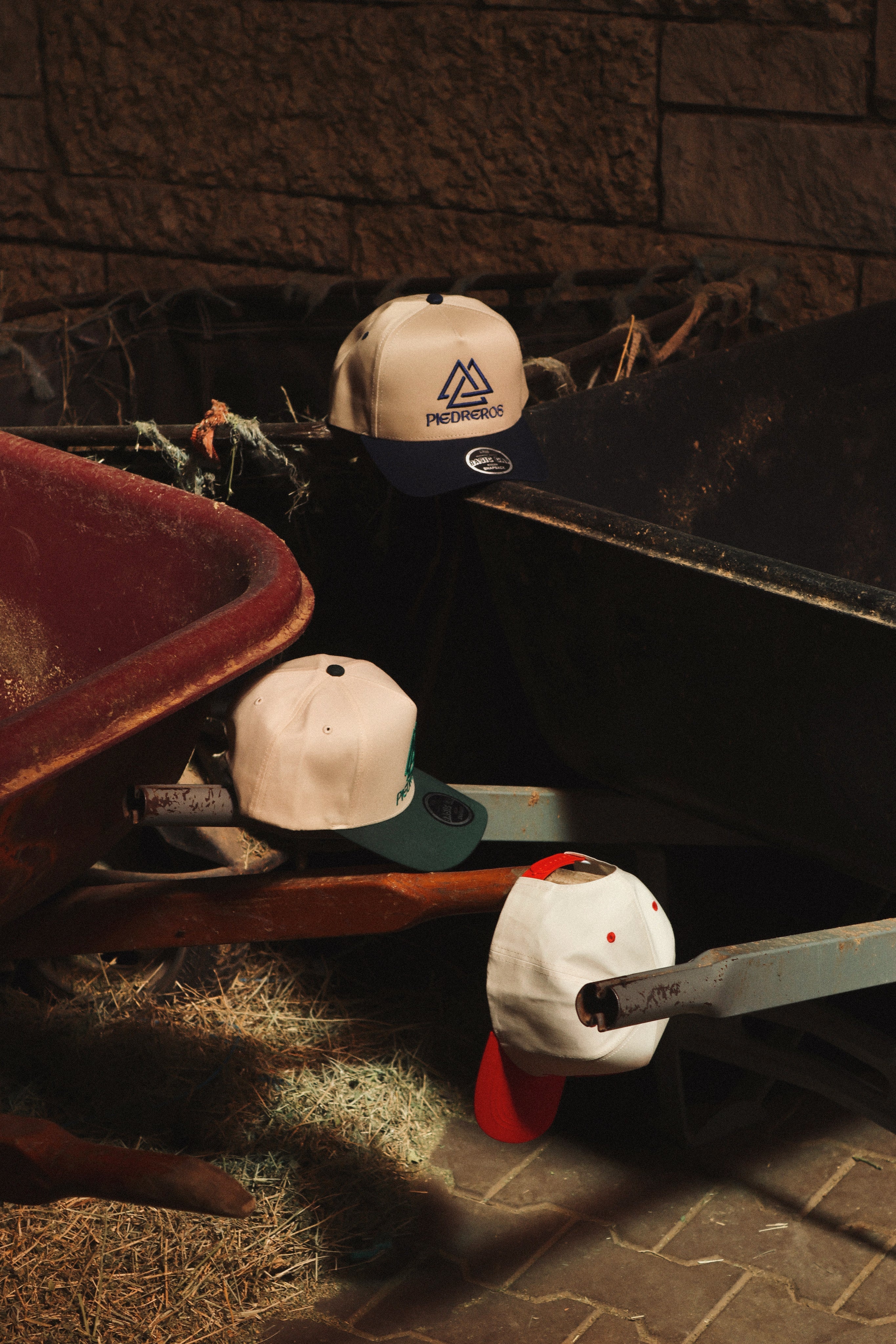 Piedreros Basic Cap en tres colorways sobre carretillas, corona crema con visera de color, wordmark PIEDREROS y logo Trinidad