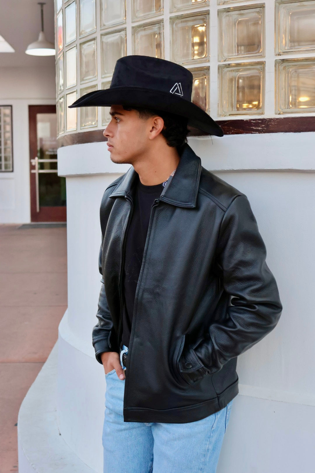Man wearing a black leather jacket and cowboy hat leaning against a building.
