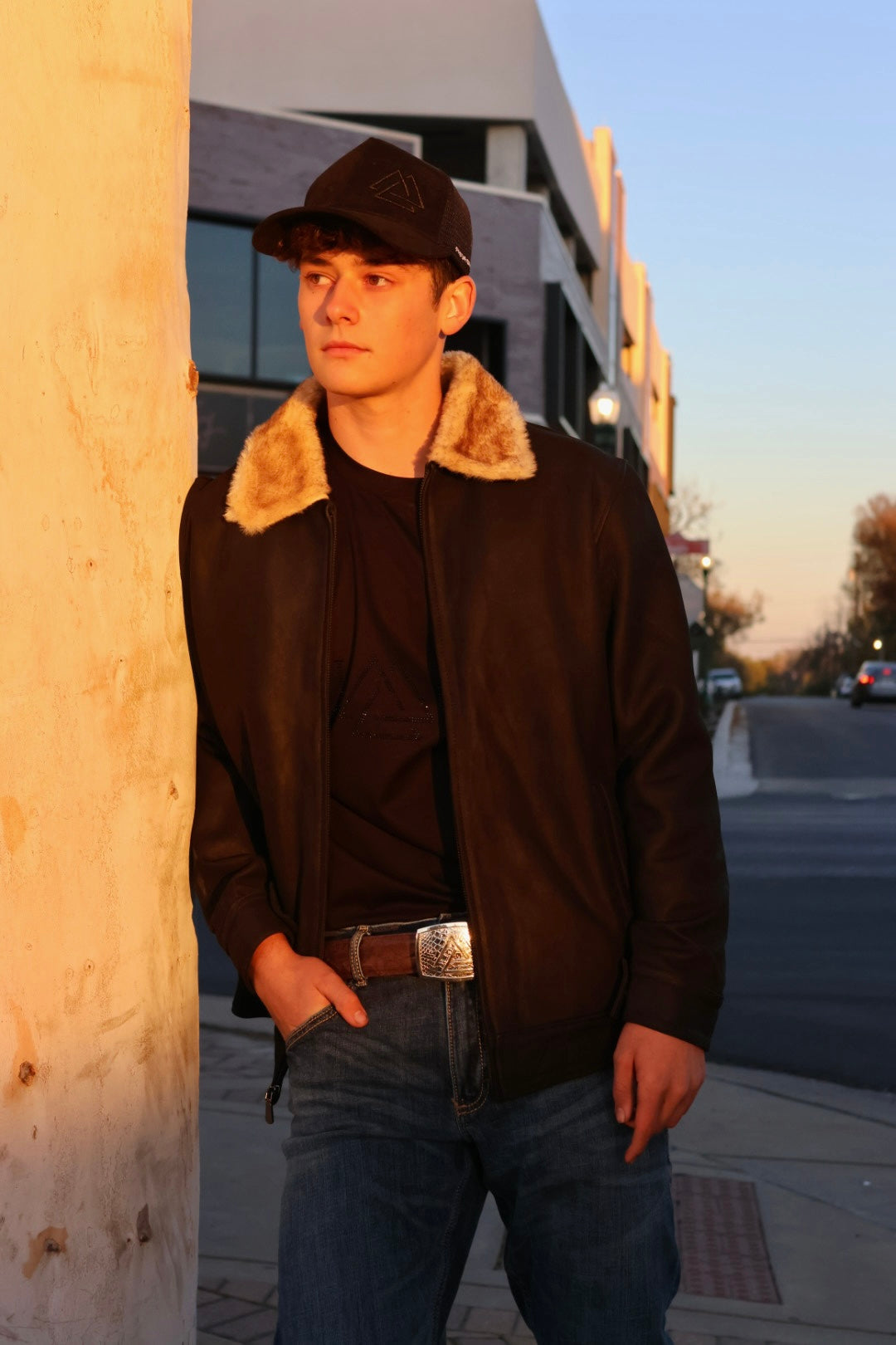 Man wearing a dark jacket with fur collar and cap leaning against a wall outdoors.