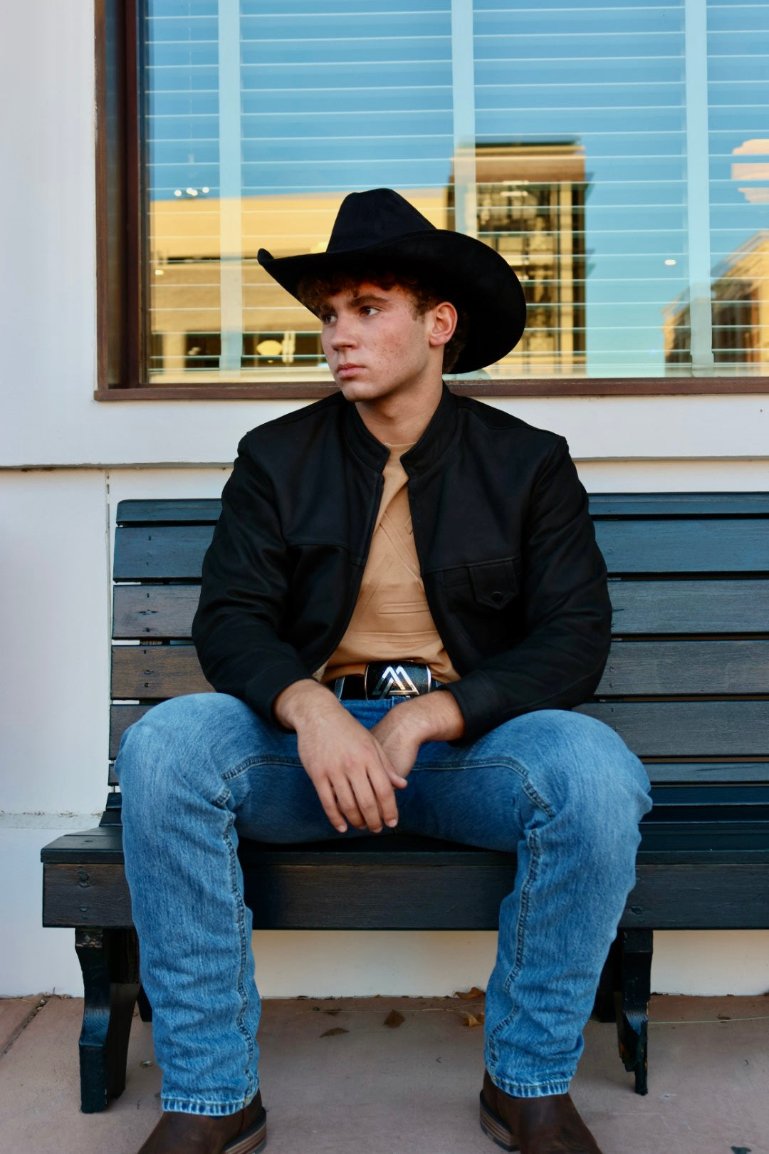 Man wearing a black cowboy hat, black jacket, beige shirt, and blue jeans sitting on a bench.