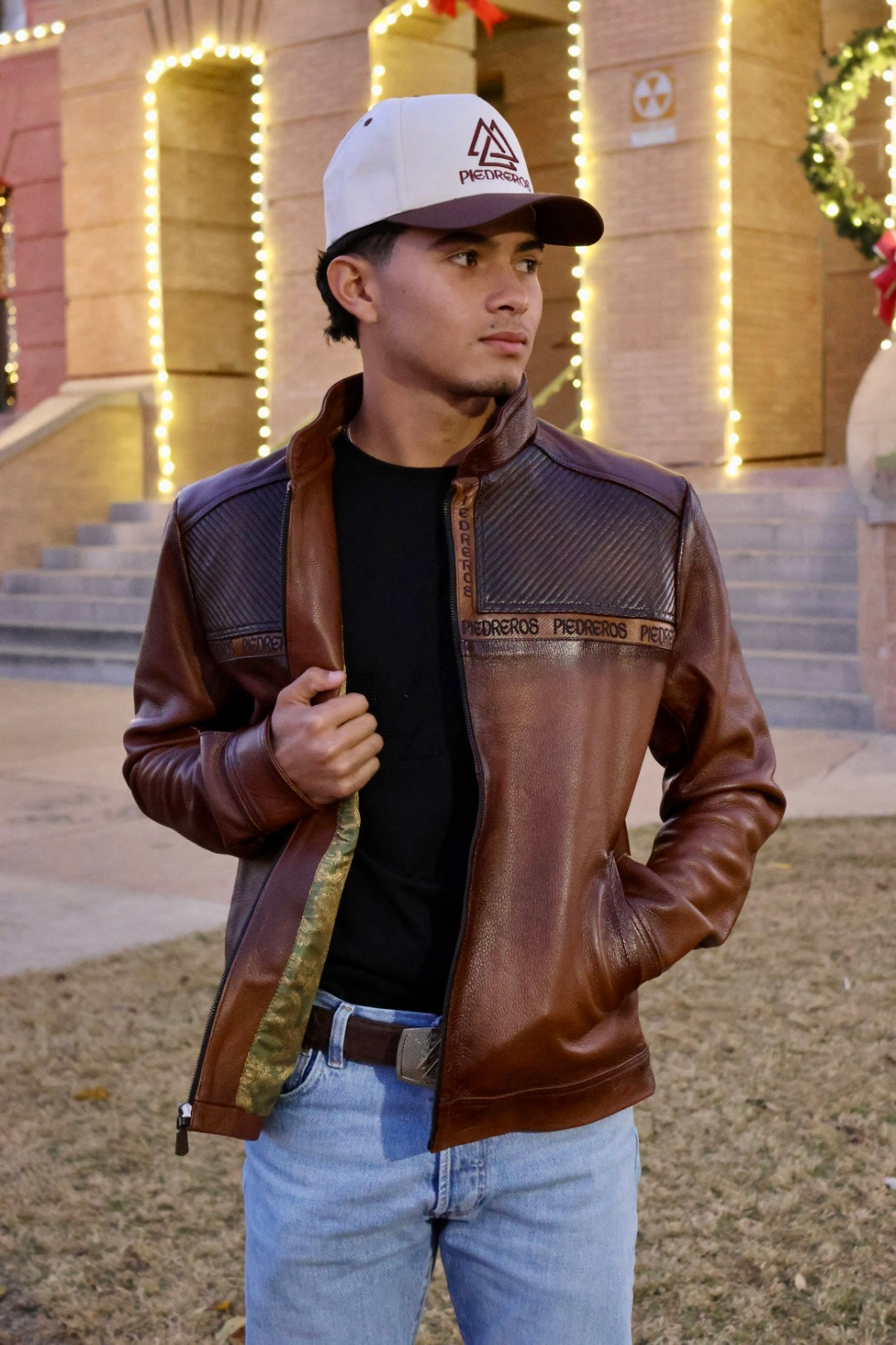 Man wearing a brown leather jacket and white cap in front of a building with decorative lights.