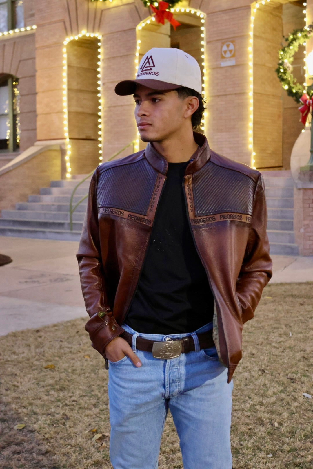 Man wearing a brown leather jacket, black shirt, and blue jeans standing outdoors with decorative lights in the background.